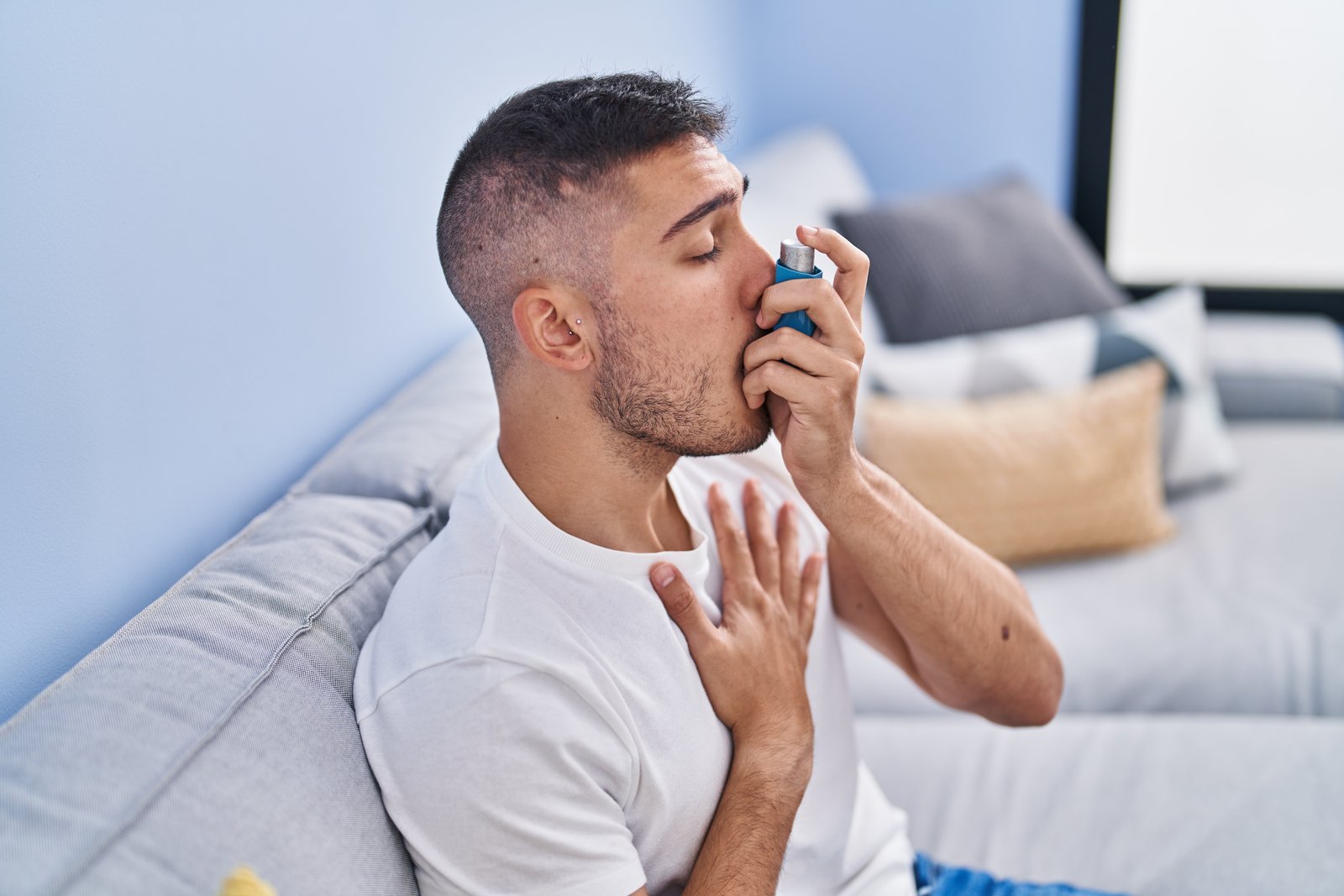 young hispanic man using inhaler sitting on sofa at home